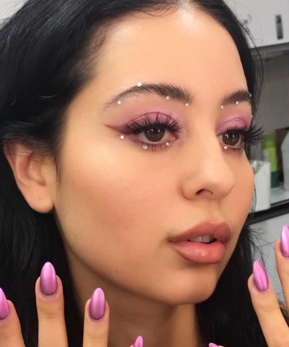 Maquiagens de euphoria com sombra rosa, delineado gráfico, pedrarias nos olhos e unhas lilás em close glamouroso