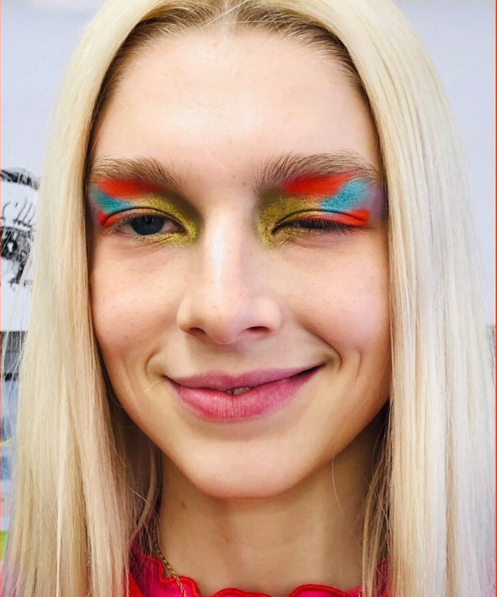 Maquiagens de euphoria em modelo sorrindo e piscando, com sombra neon azul, laranja e dourada, look vibrante e ousado