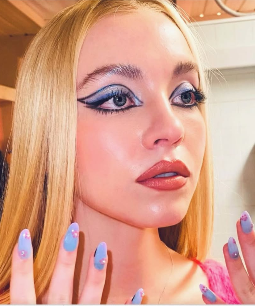 Maquiagens de euphoria com delineado gráfico azul, brilho prateado, unhas pastel e modelo posa com olhar intenso.