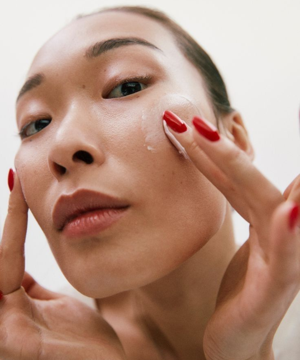 Protocolos de beleza em close: modelo aplica creme no rosto com unhas vermelhas, pele luminosa e clima de autocuidado.