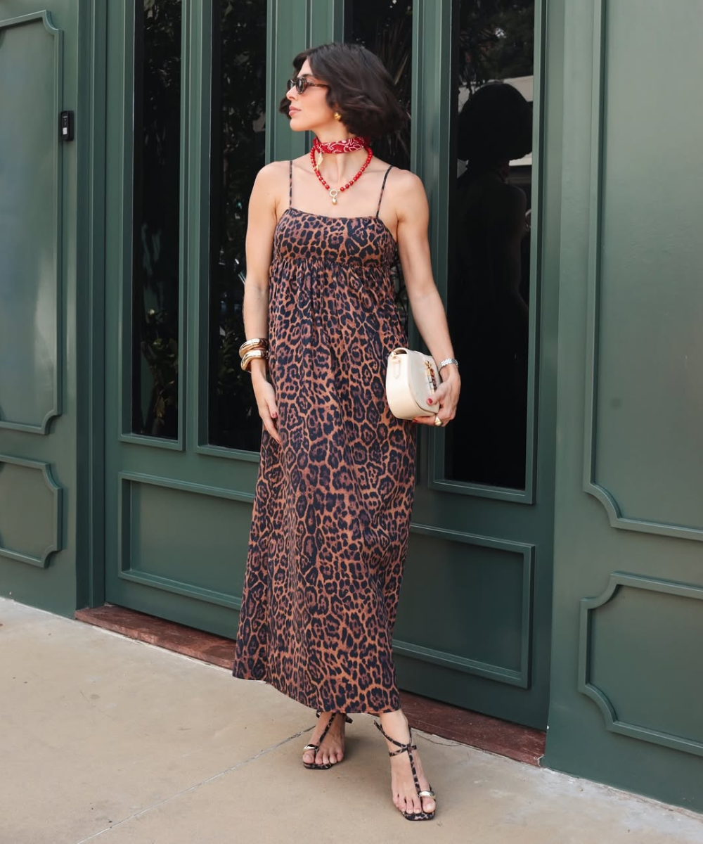 Modelo posa com lenço com pingente vermelho, vestido longo animal print e bolsa clara diante de fachada verde elegante.