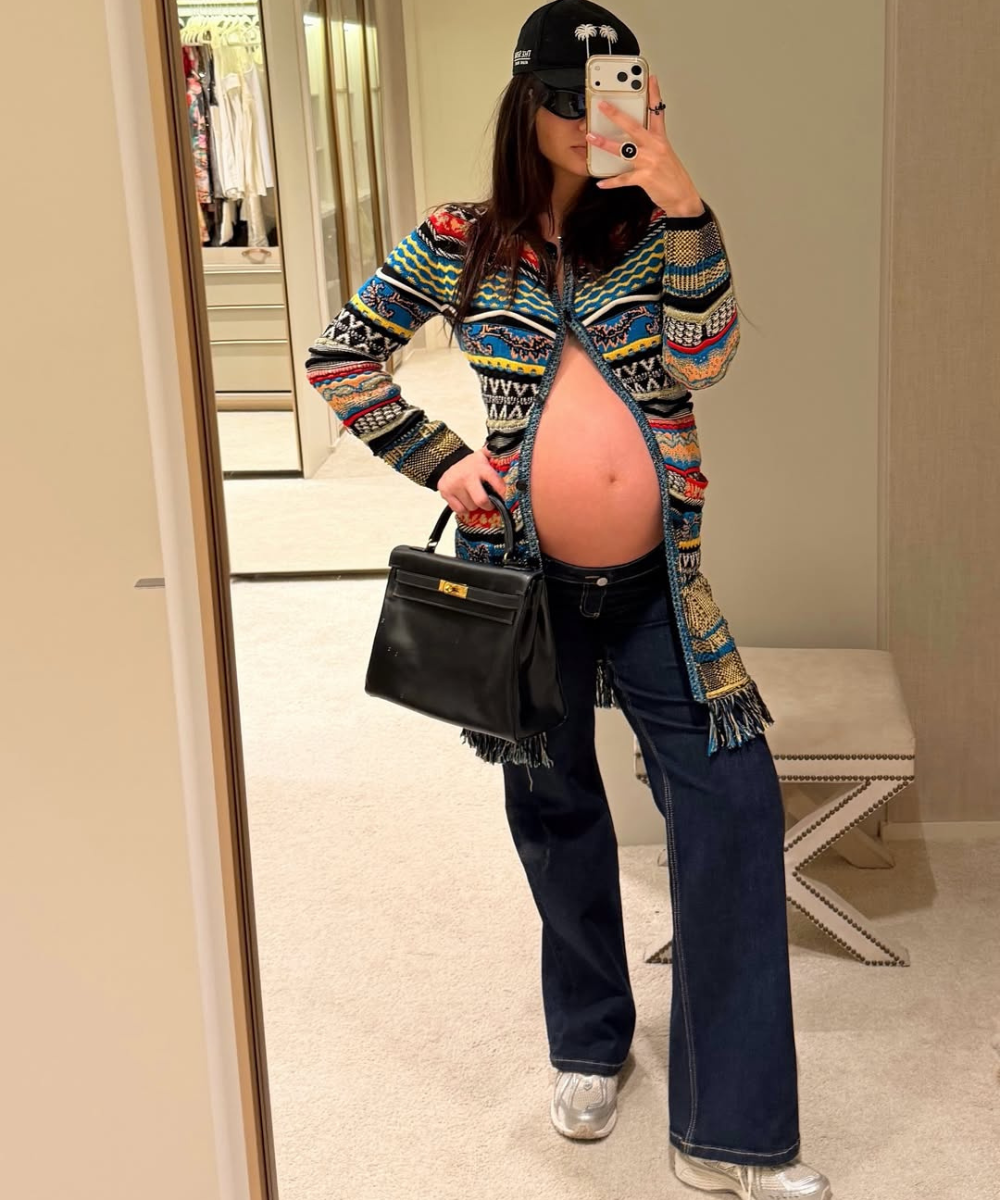 Looks de grávidas com modelo posa no espelho em tricô estampado, jeans flare e boné preto, clima despojado.