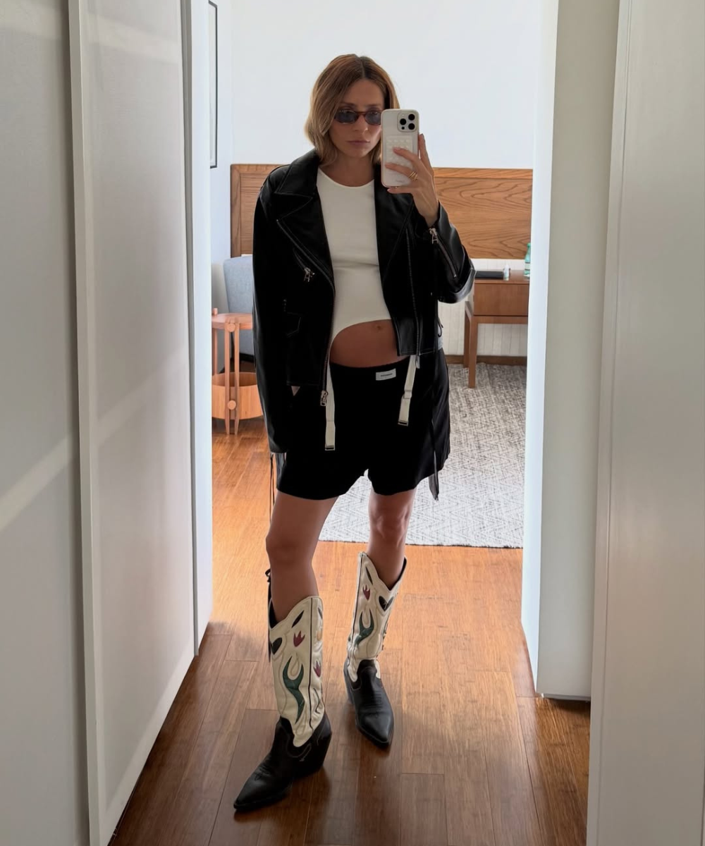 Looks de grávidas com jaqueta de couro preta, top branco, bermuda preta e bota western; modelo posa com atitude em espelho.