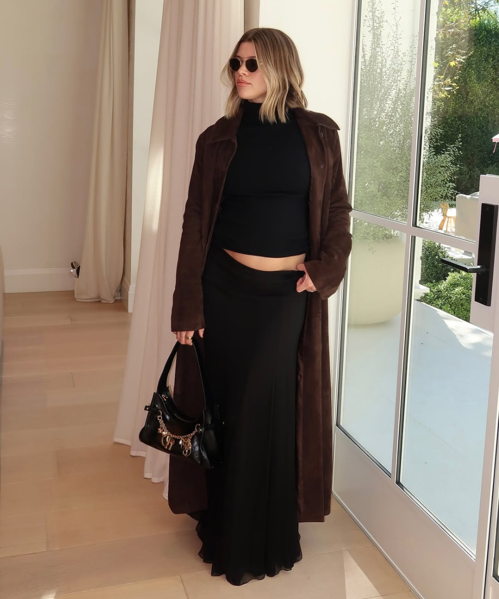 Looks de grávidas com modelo posa em conjunto preto ajustado, casaco marrom de camurça e bolsa preta, clima elegante.