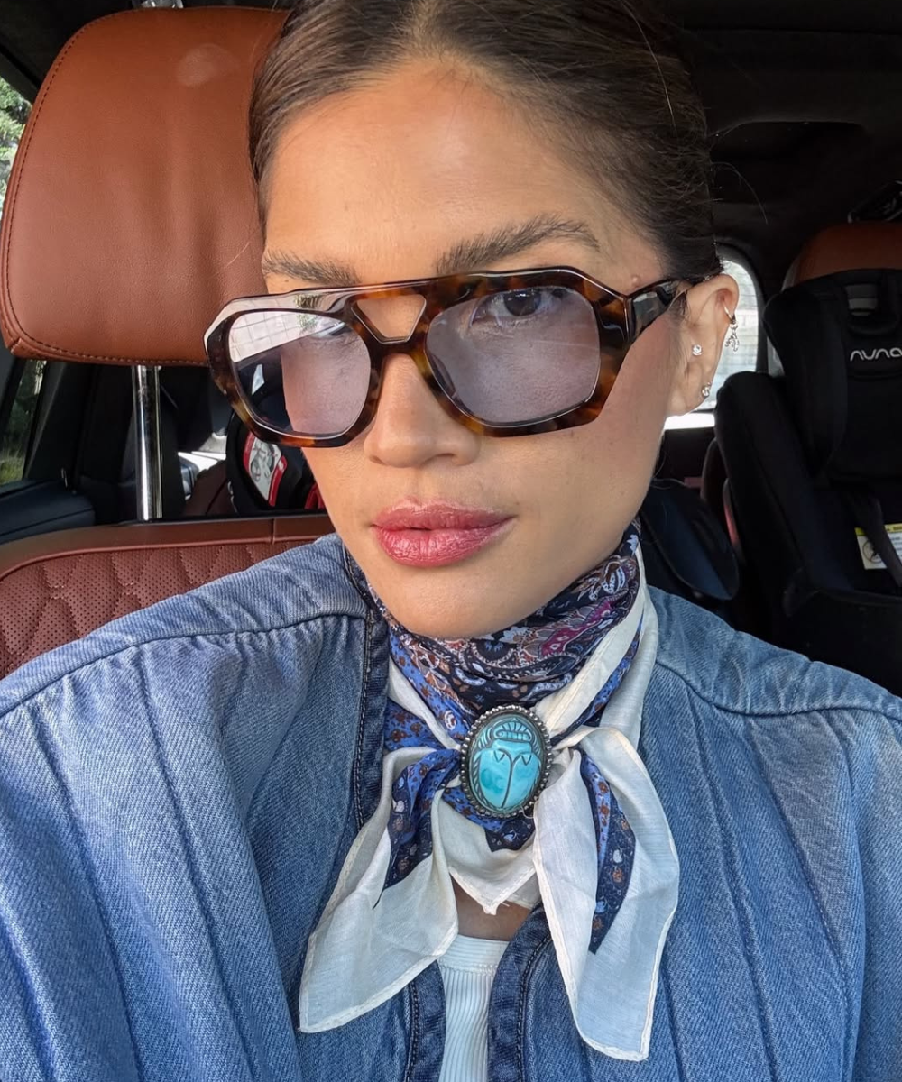 Modelo posa no carro com lenço com pingente azul, camisa jeans e óculos tartaruga, em visual elegante e sereno.