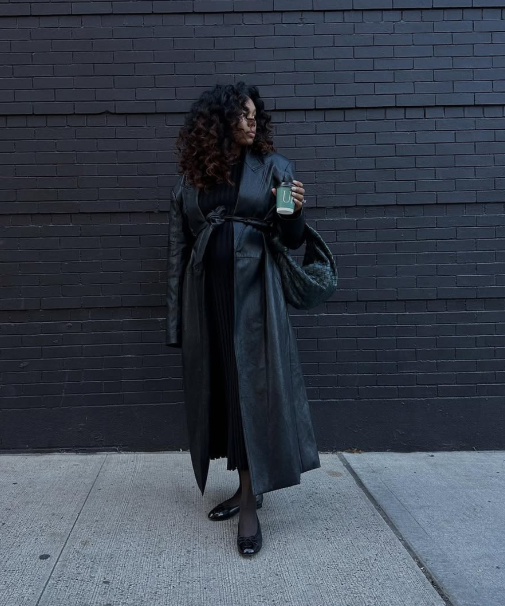 Looks de trabalho para usar durante a gravidez: modelo posa de preto com vestido plissado e trench de couro, elegante.