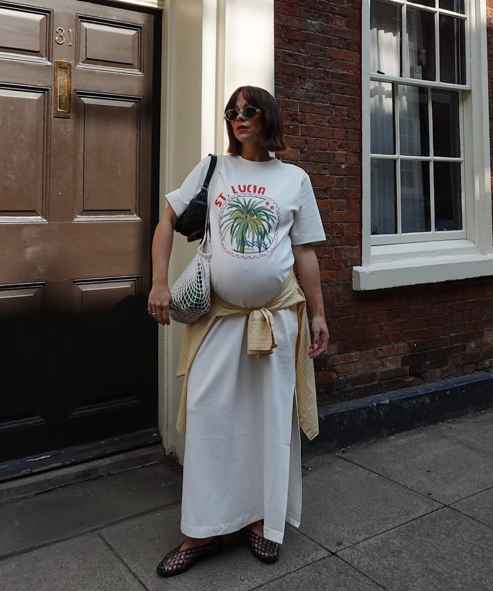 Looks de trabalho para usar durante a gravidez com vestido branco amplo; modelo posa serena na calçada. Texto: ST. LUCIA