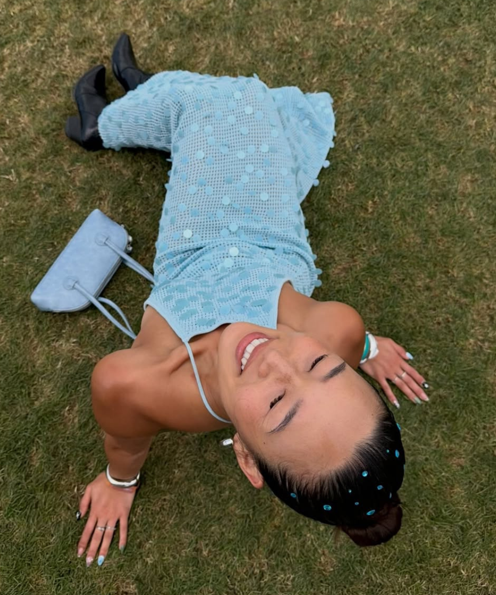 Modelo posa sorrindo na grama com strass no cabelo, vestido azul texturizado, bolsa combinando e botas pretas.