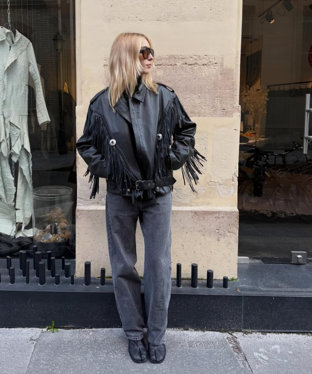 Looks com franja: modelo posa de lado com jaqueta preta de couro franjada, jeans cinza e botas, em clima urbano sofisticado.