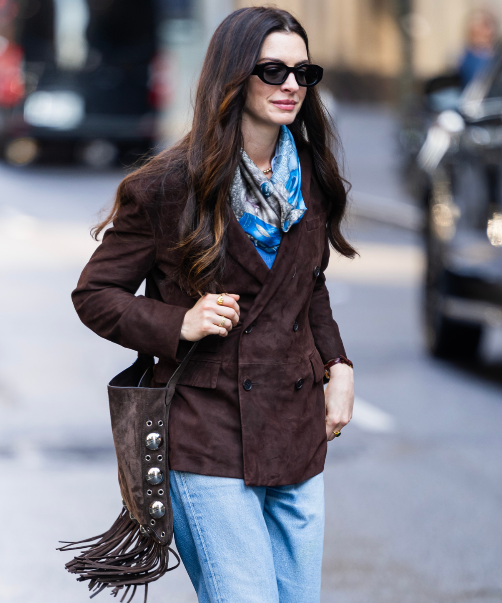 Looks com franja: modelo posa ao caminhar com blazer marrom, jeans claros, lenço azul e bolsa de camurça boho elegante.