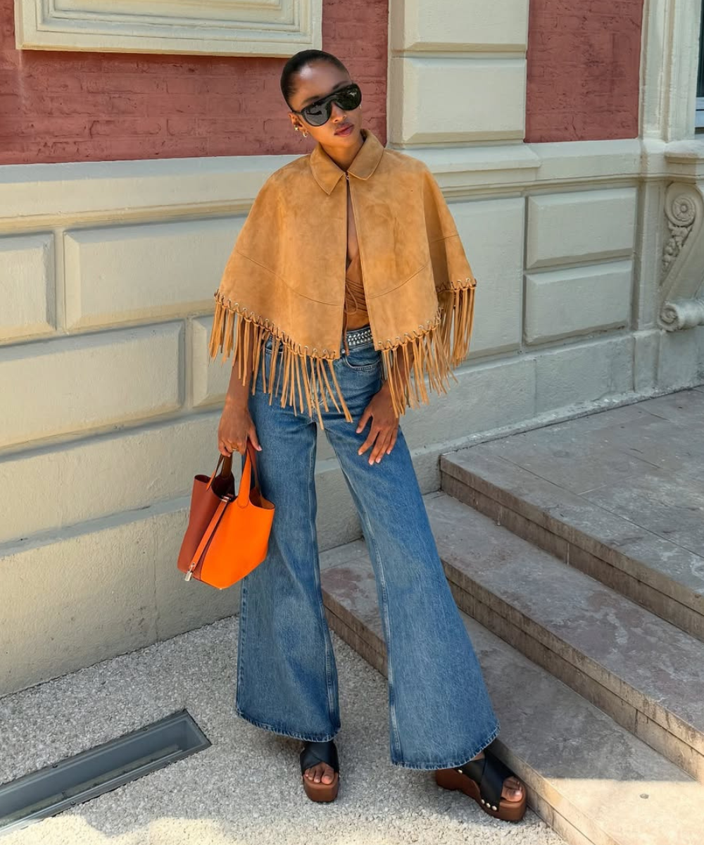 Looks com franja: modelo posa com capa camel de camurça, jeans amplo azul e bolsa laranja em clima sofisticado.