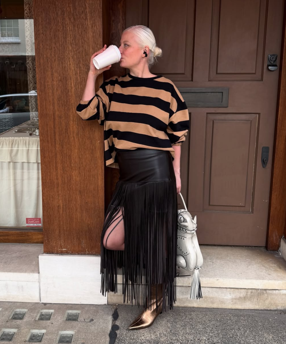 Looks com franja: modelo posa com blusa listrada bege e preta, saia preta com franjas e bota metalizada, clima urbano