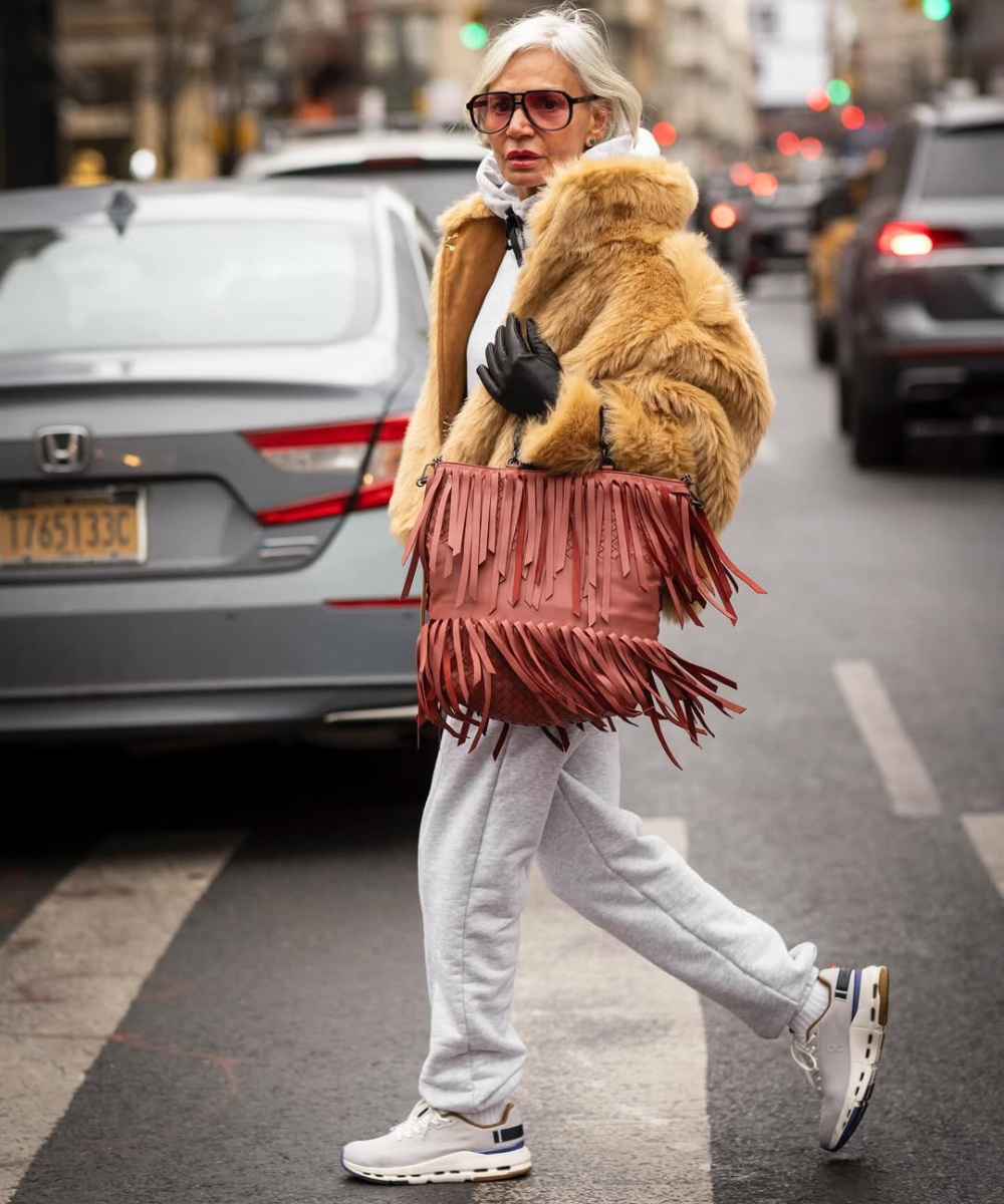 Looks com franja: modelo cruza a rua com casaco de pele caramelo, moletom cinza e bolsa marrom, em clima urbano elegante.