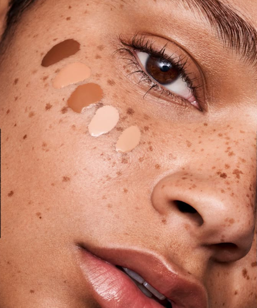 Close no rosto com sardas e amostras de corretivo mais claro que a base, em tons bege, com pele luminosa e olhar suave