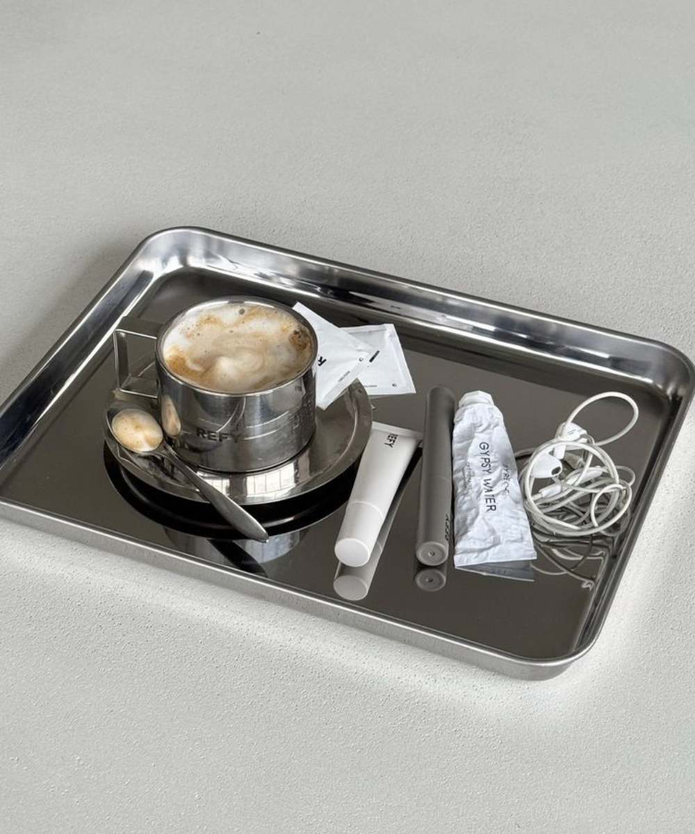 Bandeja metálica espelhada com café cremoso, tubos minimalistas e fones brancos em cenário cinza sereno.