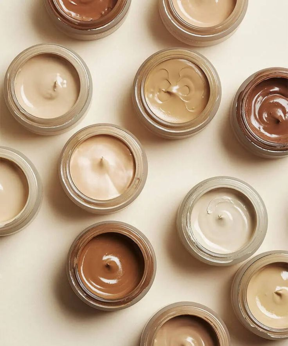 Corretivo tem que ser mais claro que a base em potes cremosos bege e marrom sobre fundo claro, com clima clean e elegante.