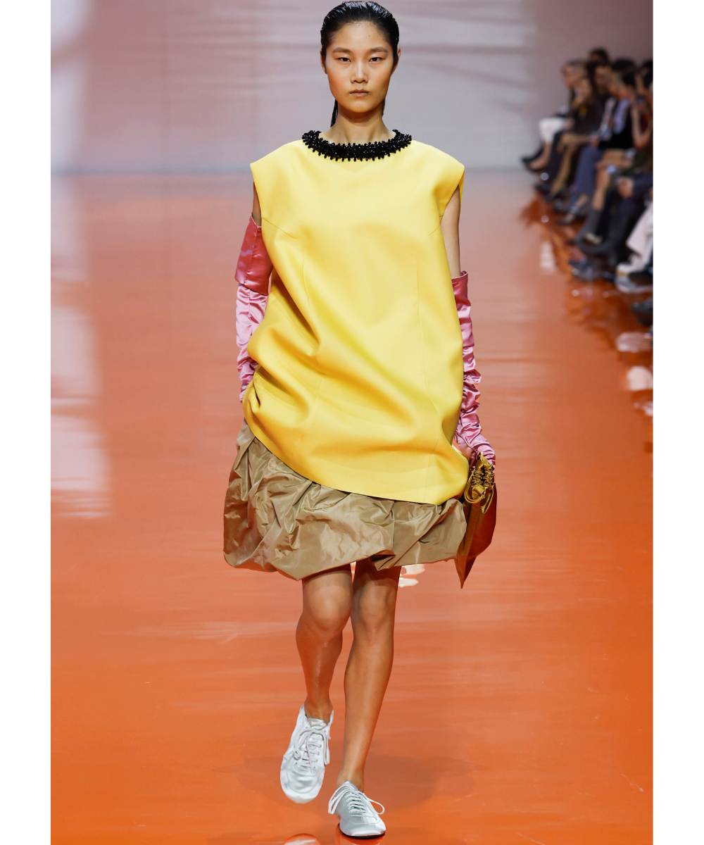 Modelo desfila com vestido amarelo amplo, mangas rosa acetinadas, saia drapeada e sapatos de cetim brancos em clima ousado.