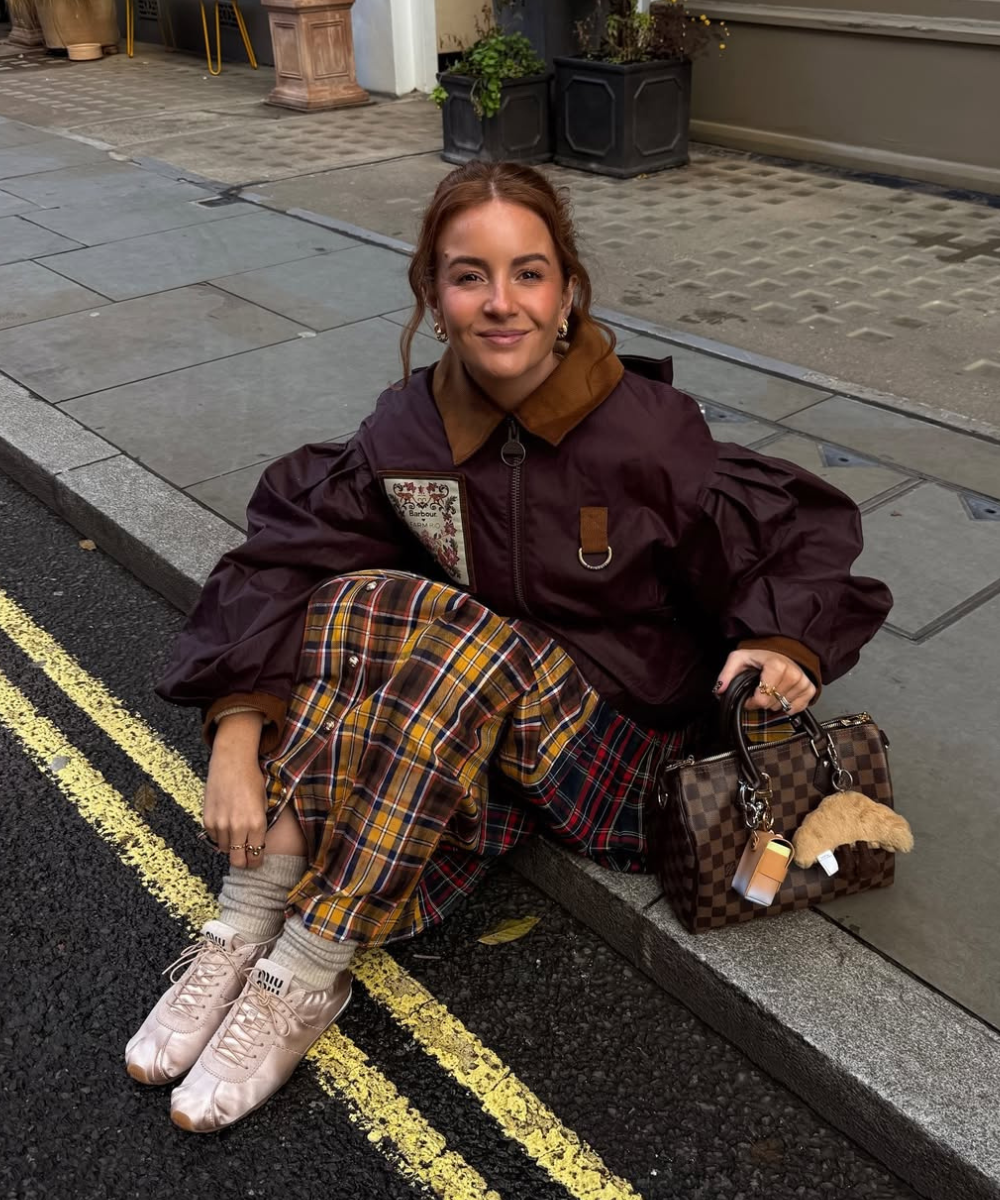 Modelo posa na calçada com sapatos de cetim rosa, jaqueta vinho ampla, saia xadrez e bolsa marrom em clima casual elegante.