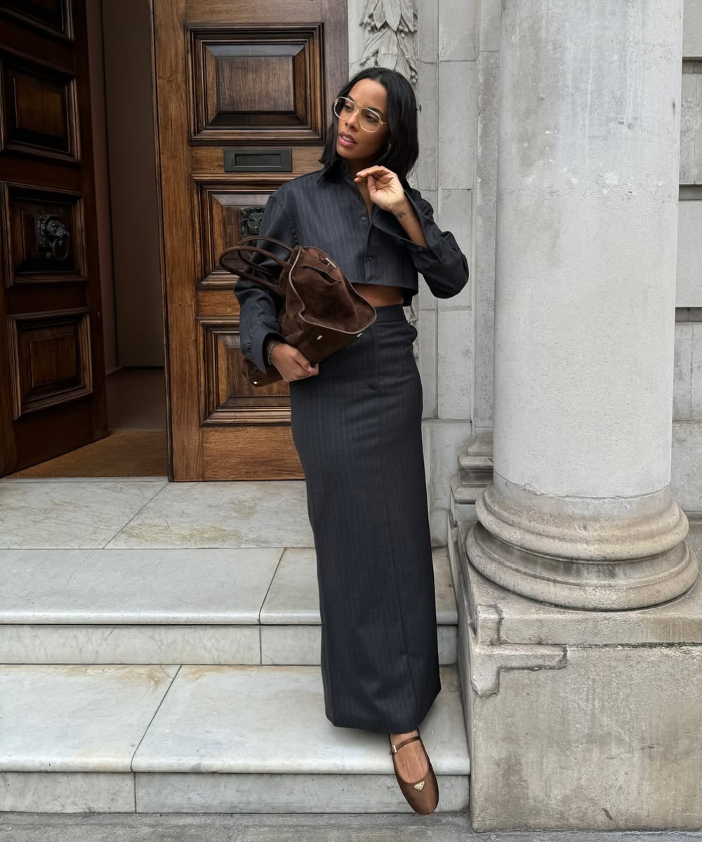 Modelo posa com conjunto risca de giz grafite, bolsa marrom e sapatos de cetim marrons diante de porta clássica, clima