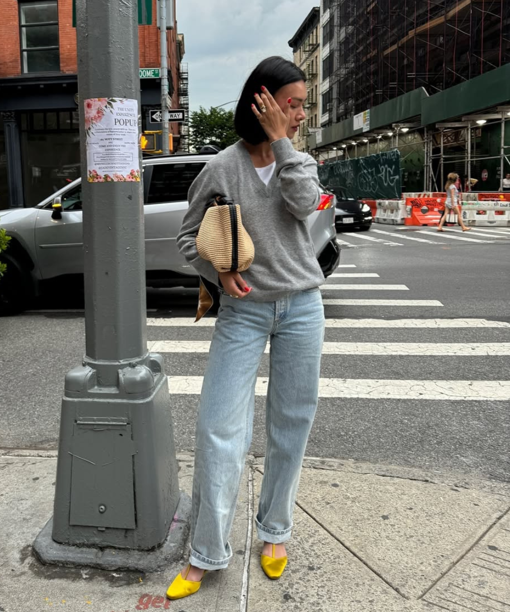 Modelo posa na esquina com suéter cinza, jeans claros e sapatos de cetim amarelos, em clima urbano elegante.