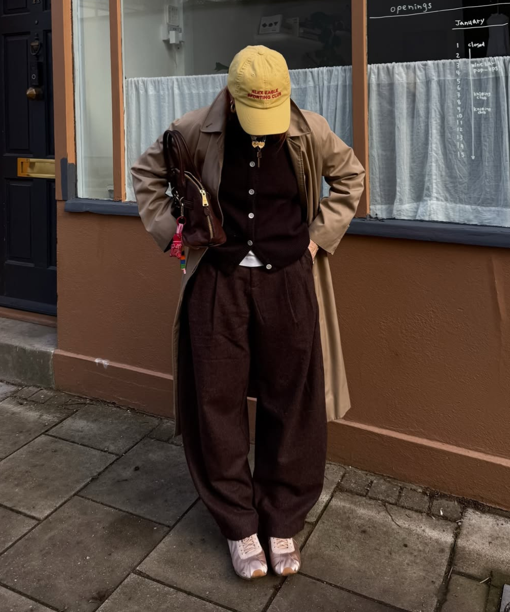 Modelo posa com sapatos de cetim rosados, trench bege e alfaiataria marrom em clima urbano casual.