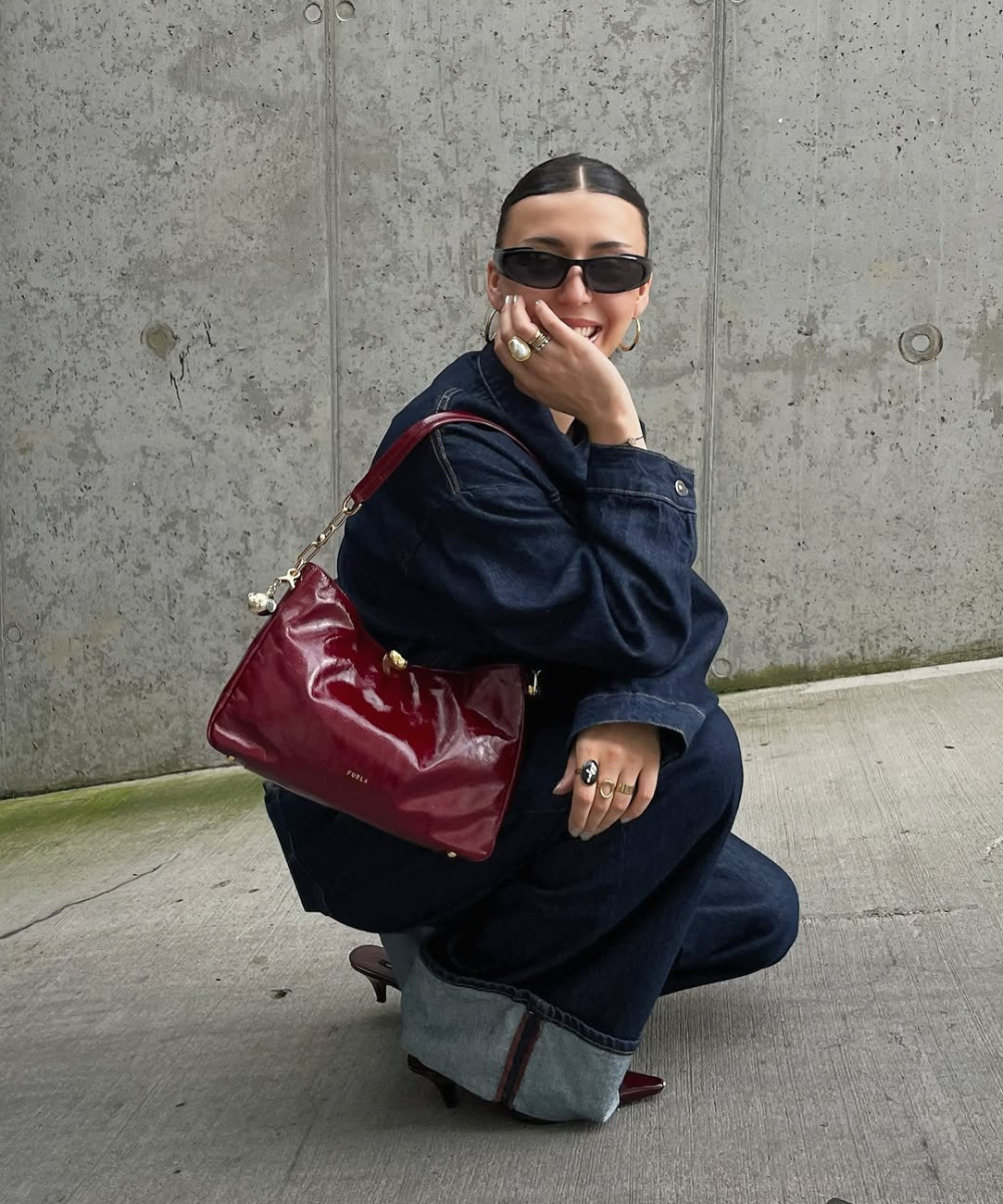Modelo posa agachada com jeans amplo, bolsa vinho envernizada chic para o outono e óculos escuros diante do concreto.