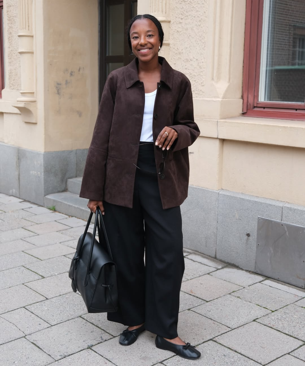 Modelo posa com jaqueta marrom de camurça, calça ampla preta e bolsa preta, uma das cores de bolsa mais chiques para o