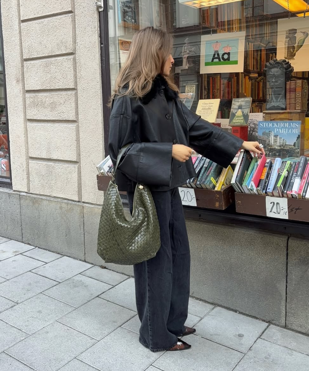 Cores de bolsa mais chiques para o outono: modelo posa com bolsa verde-oliva tramada, jaqueta preta e clima urbano culto.
