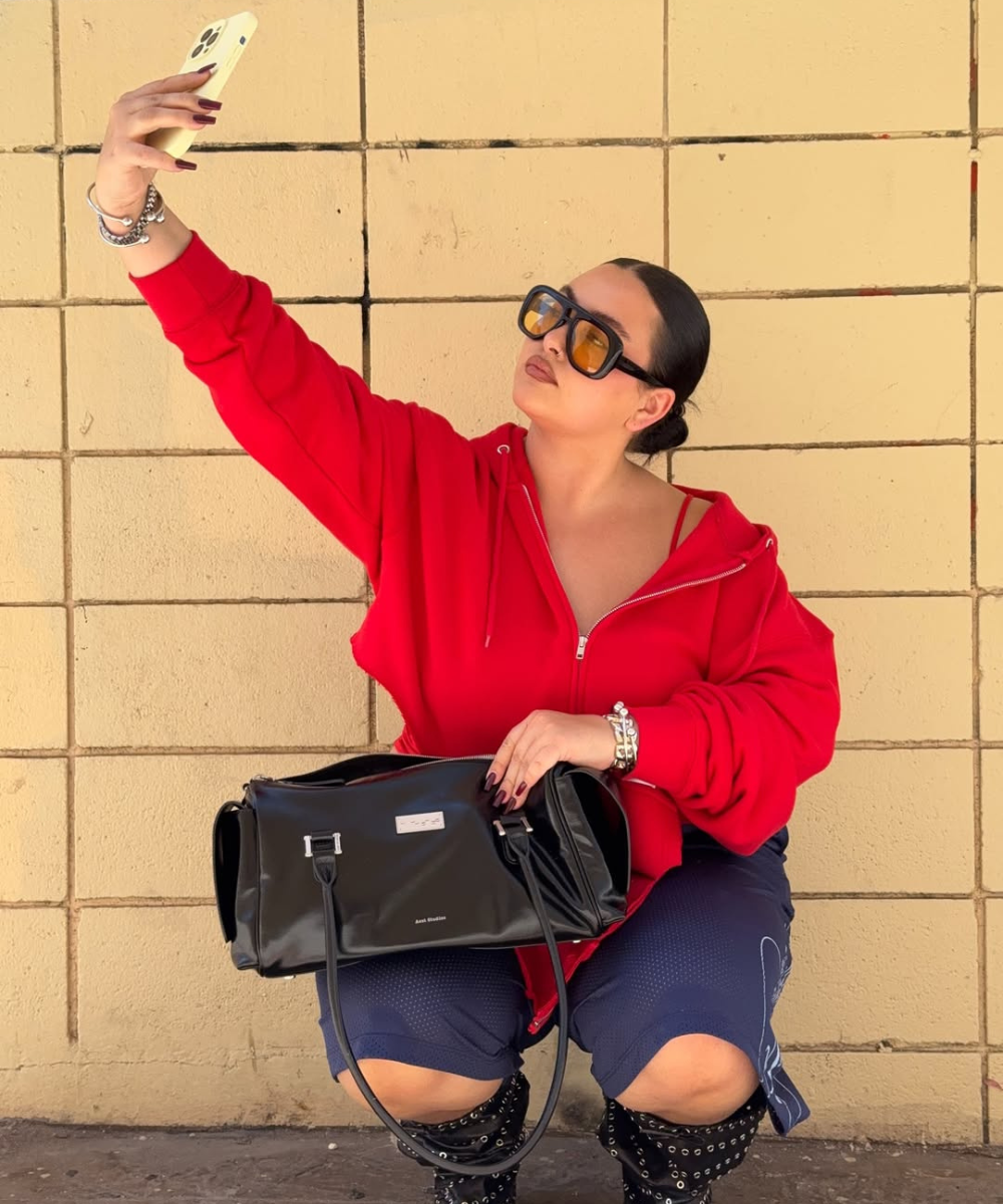 Modelo posa agachada com moletom vermelho, óculos maxi âmbar e bolsa preta brilhante, acessórios para o outono urbanos.