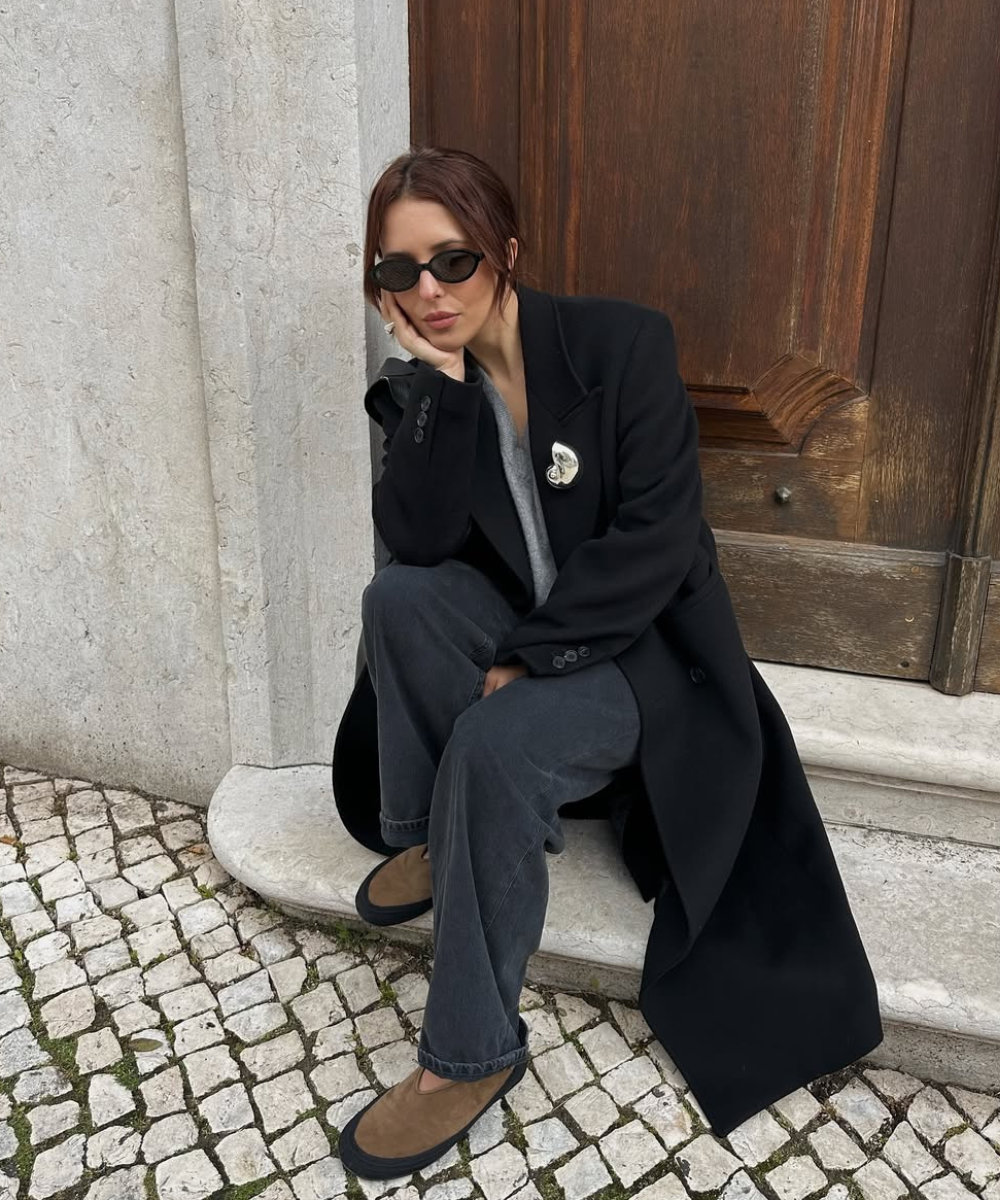 Acessórios para o outono: modelo posa sentada com óculos pretos, broche metálico, sobretudo preto e tons sóbrios elegantes