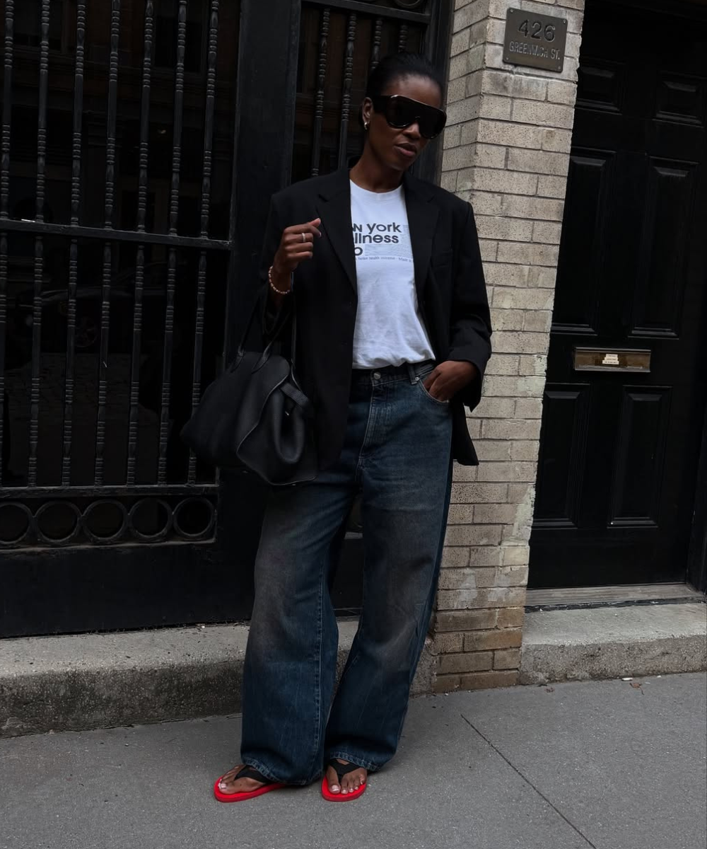 Acessórios para o outono: modelo posa de blazer preto, jeans amplo, bolsa e óculos escuros; chinelo vermelho.