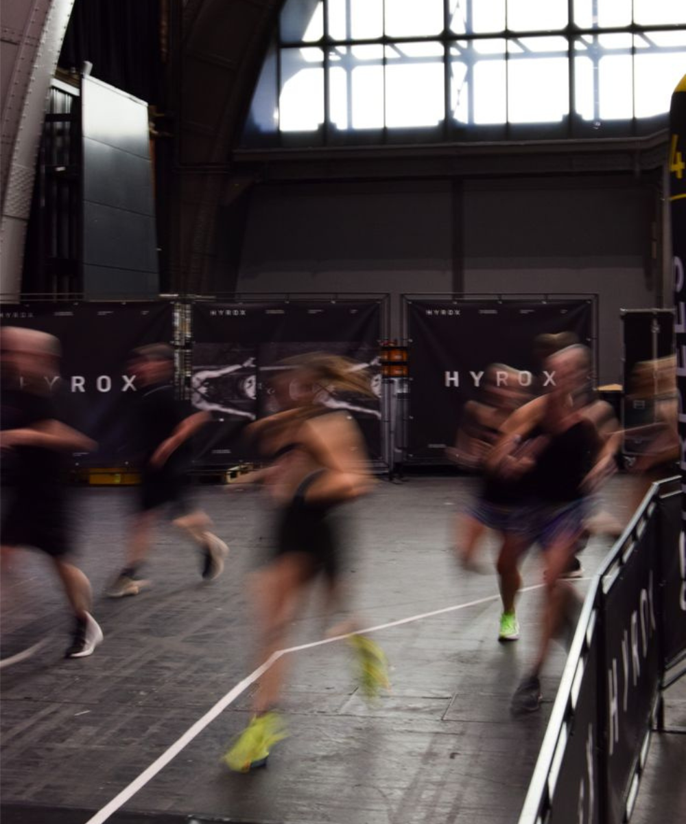 Atletas correm em prova Hyrox com looks esportivos pretos e tênis neon, em arena escura de clima intenso; Texto: HYROX