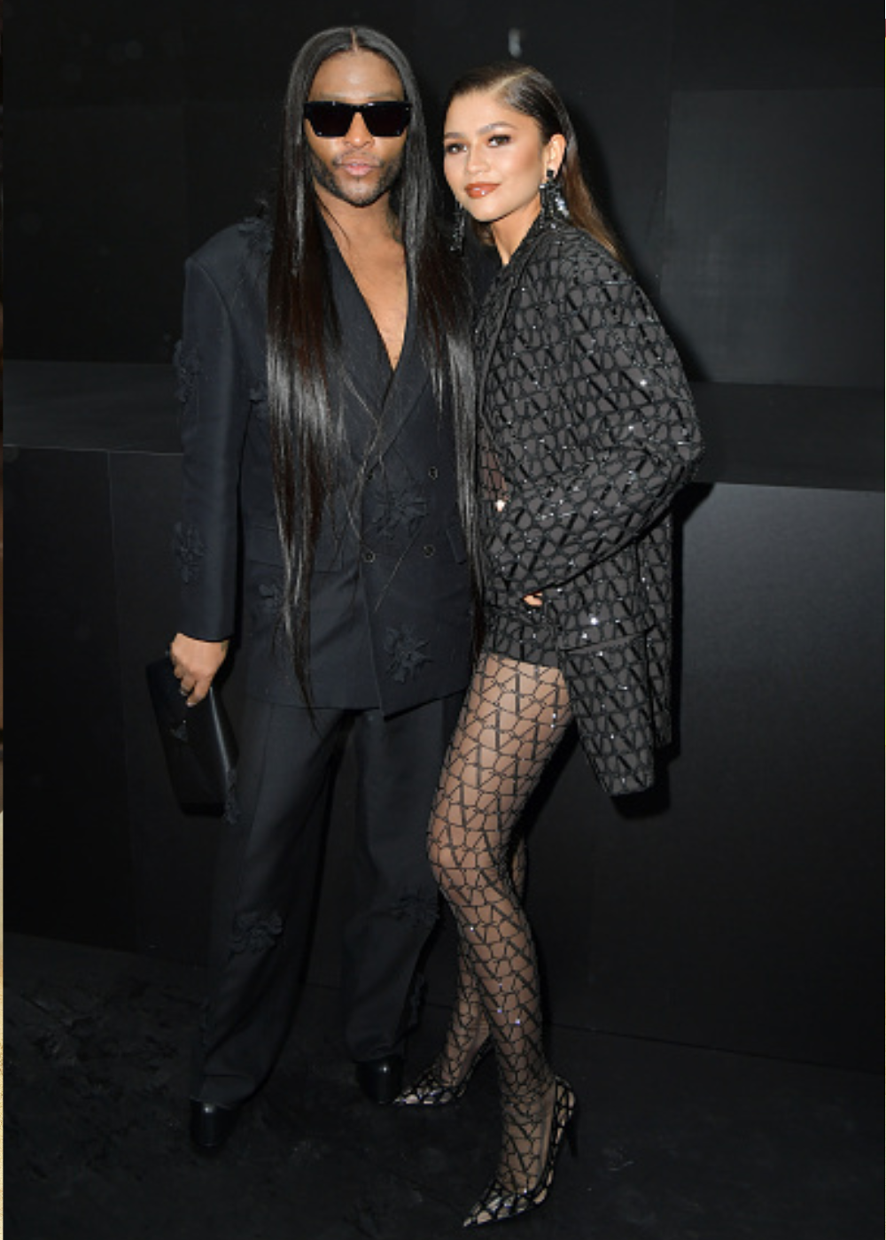 Looks da Zendaya em preto: atriz posa com blazer texturizado, meia geométrica e salto, ao lado de homem de terno amplo.