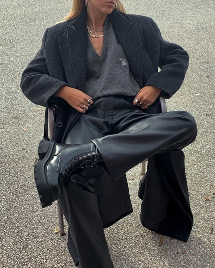 Looks com calça de couro preta wide leg, modelo posa sentada com blazer escuro, tricô cinza e coturno robusto em clima
