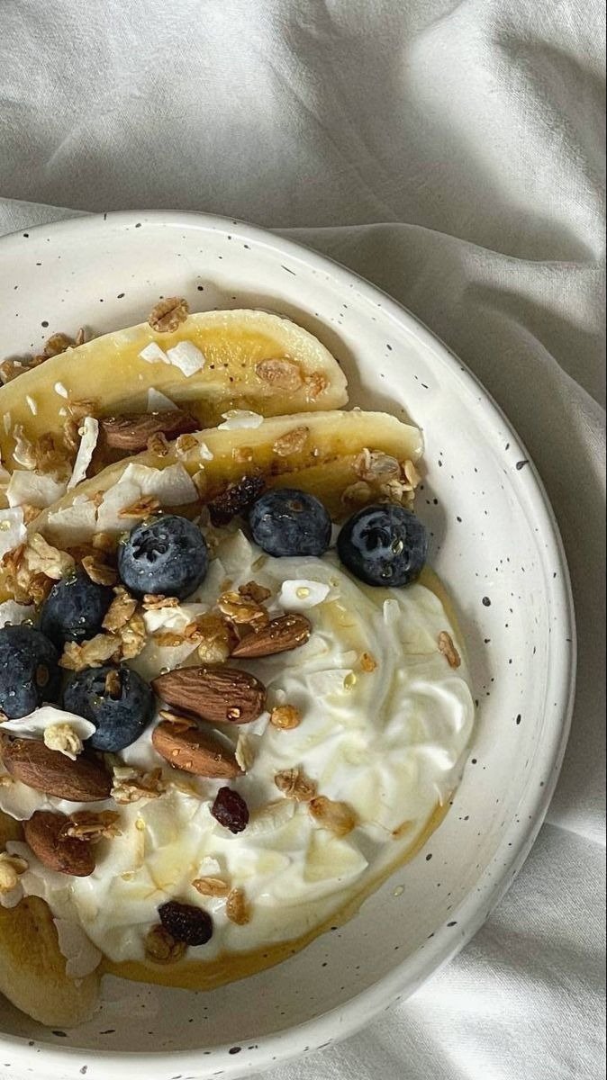 Receitas com iogurte em tigela de cerâmica com banana, mirtilos, granola e mel sobre tecido claro, clima acolhedor.
