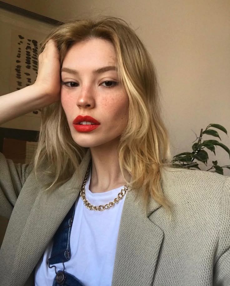 Modelo posa com blazer bege, camiseta branca e corrente dourada; cores de batom vermelho destacam beleza serena.