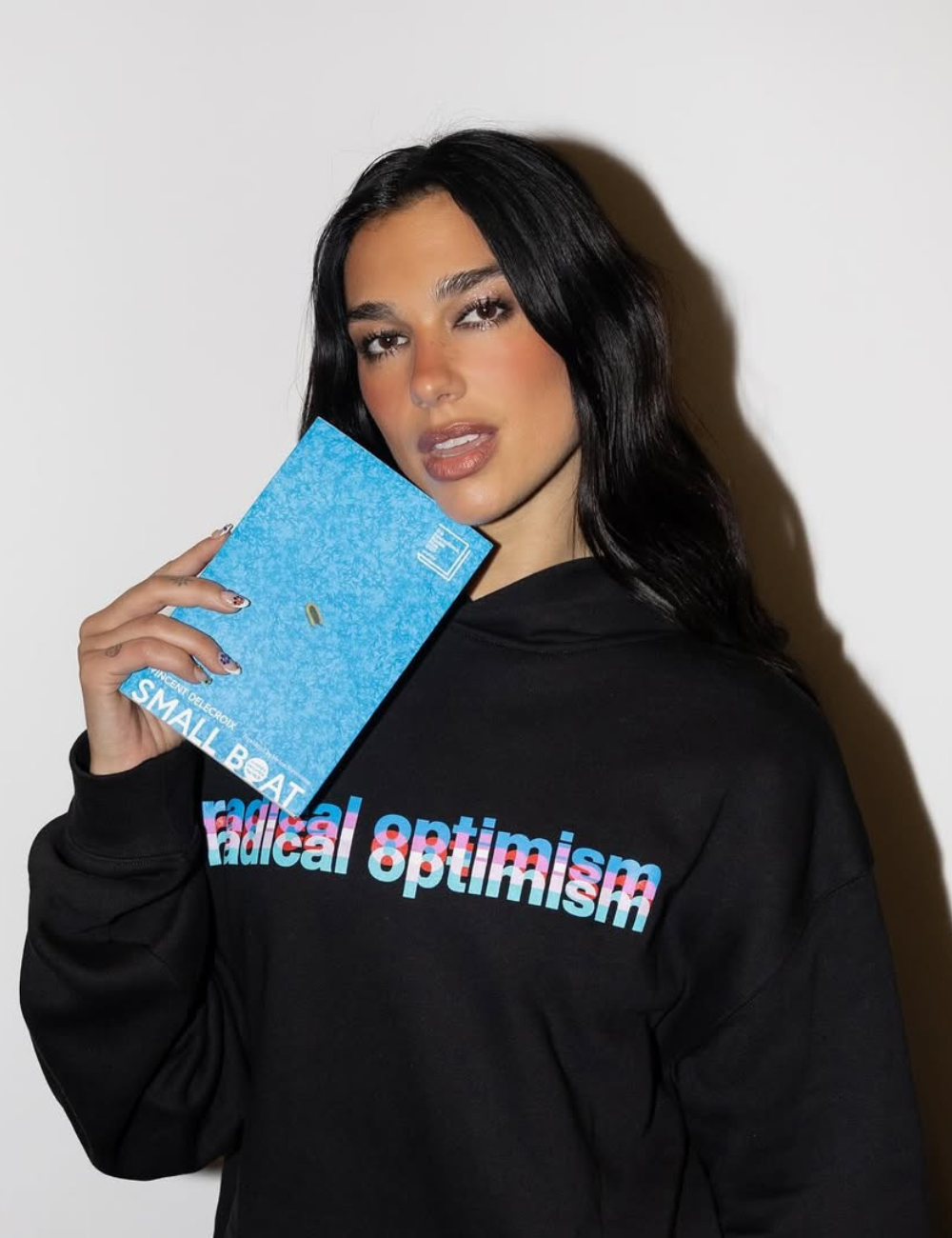 Dua Lipa posa de moletom preto oversized com livro azul. Texto: radical optimism; SMALL BOAT; VINCENT DELECROIX.