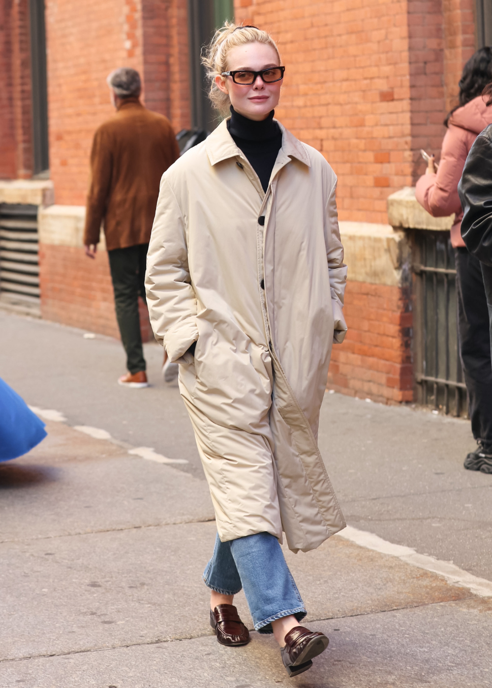 Elle Fanning caminha com elegância em casaco bege acolchoado, gola alta preta, jeans azul e mocassins vinho na rua urbana.