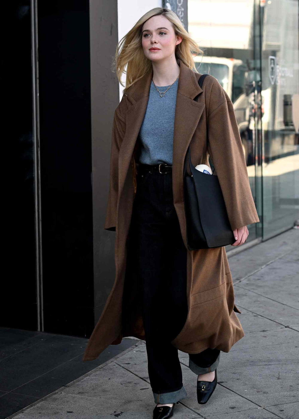 Elle Fanning caminha com elegância em sobretudo marrom amplo, malha cinza, jeans escuro e bolsa preta.