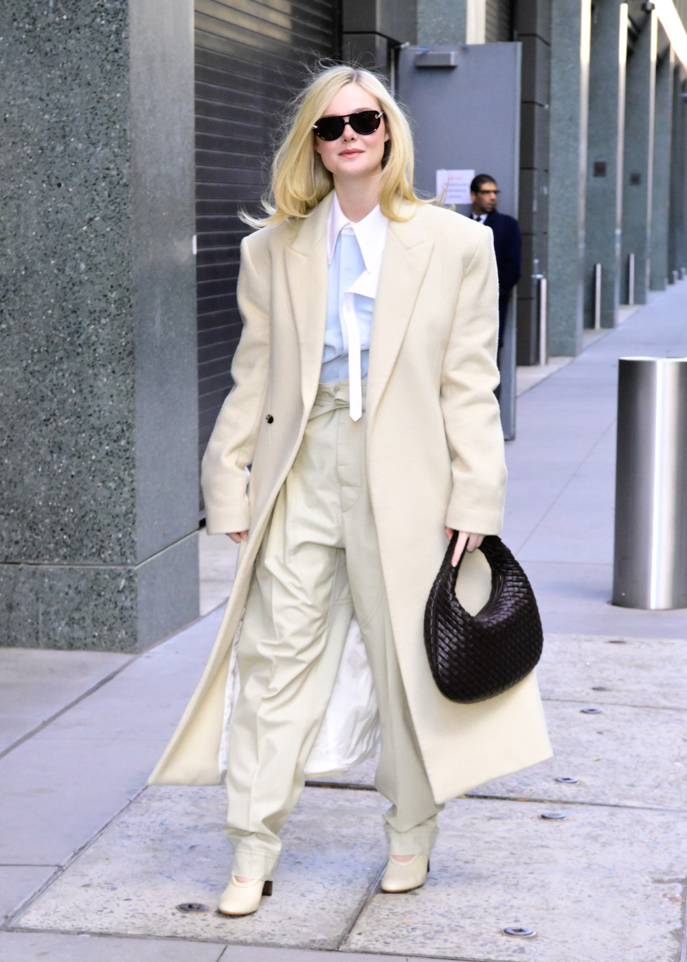 Elle Fanning posa com alfaiataria creme oversized, camisa azul-clara, óculos escuros e bolsa preta em clima elegante urbano
