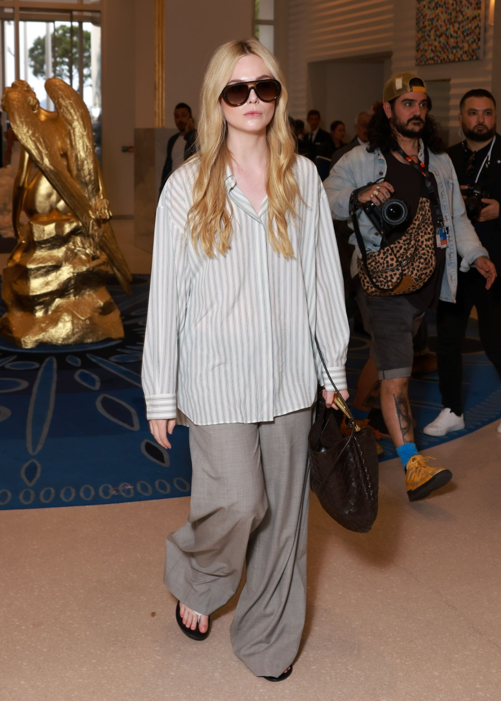 Elle Fanning caminha com camisa listrada ampla, calça cinza fluida, óculos escuros e bolsa marrom em clima sofisticado.