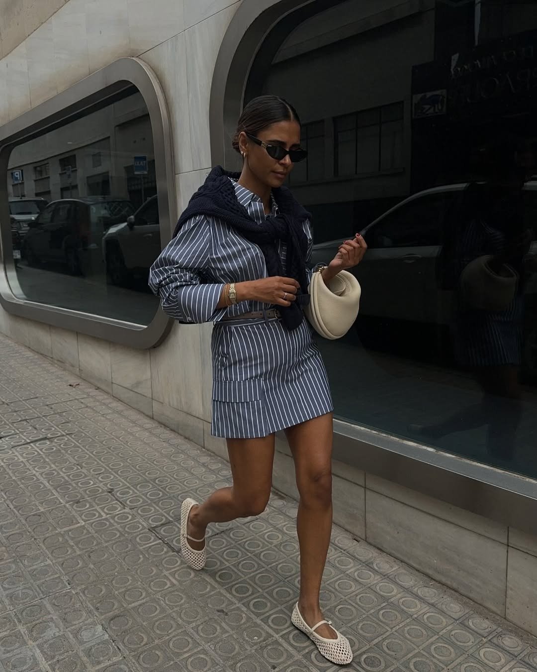 Modelo caminha com sapatilha de tela off-white, vestido listrado cinza e bolsa clara em cenário urbano elegante.