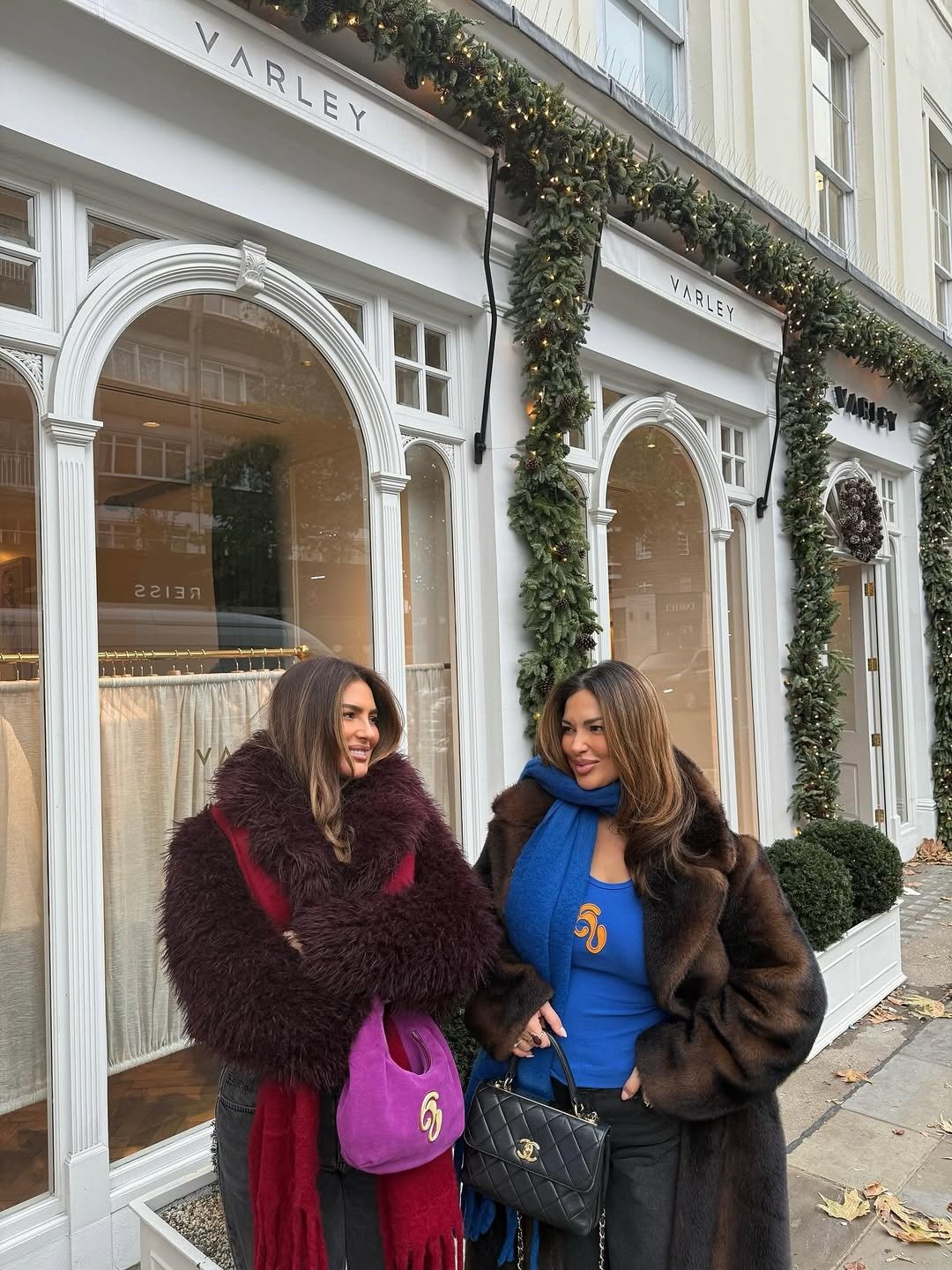 Amigas posam sorrindo com casacos felpudos vinho e marrom, azul vibrante e bolsas de luxo diante da VARLEY.