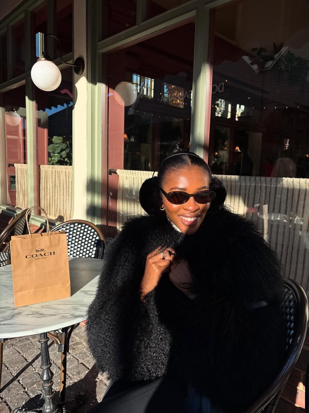 Estamos todos gostando das mesmas coisas: modelo posa de casaco felpudo preto ao sol em café. Texto: COACH NEW YORK