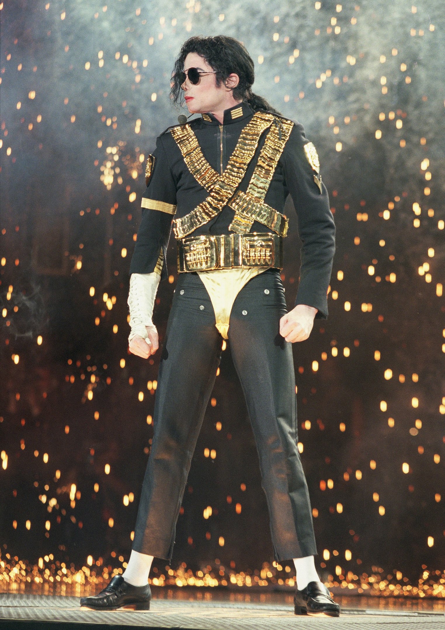 Michael Jackson posa em palco com jaqueta militar preta e dourada, calça slim e faíscas ao fundo, em clima poderoso.