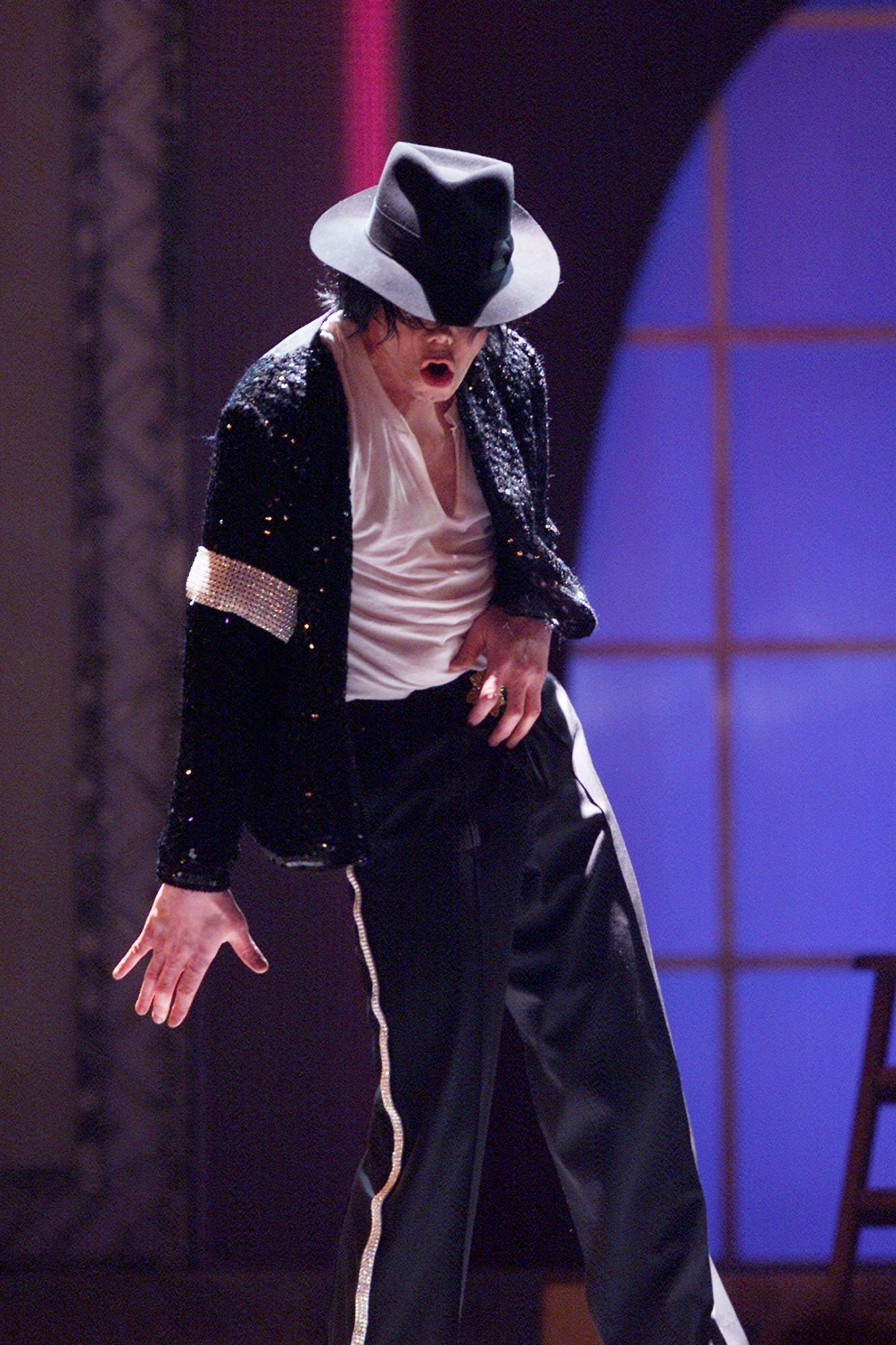 Michael Jackson dança no palco com fedora preto, blazer de paetês e camisa branca, em clima dramático e icônico