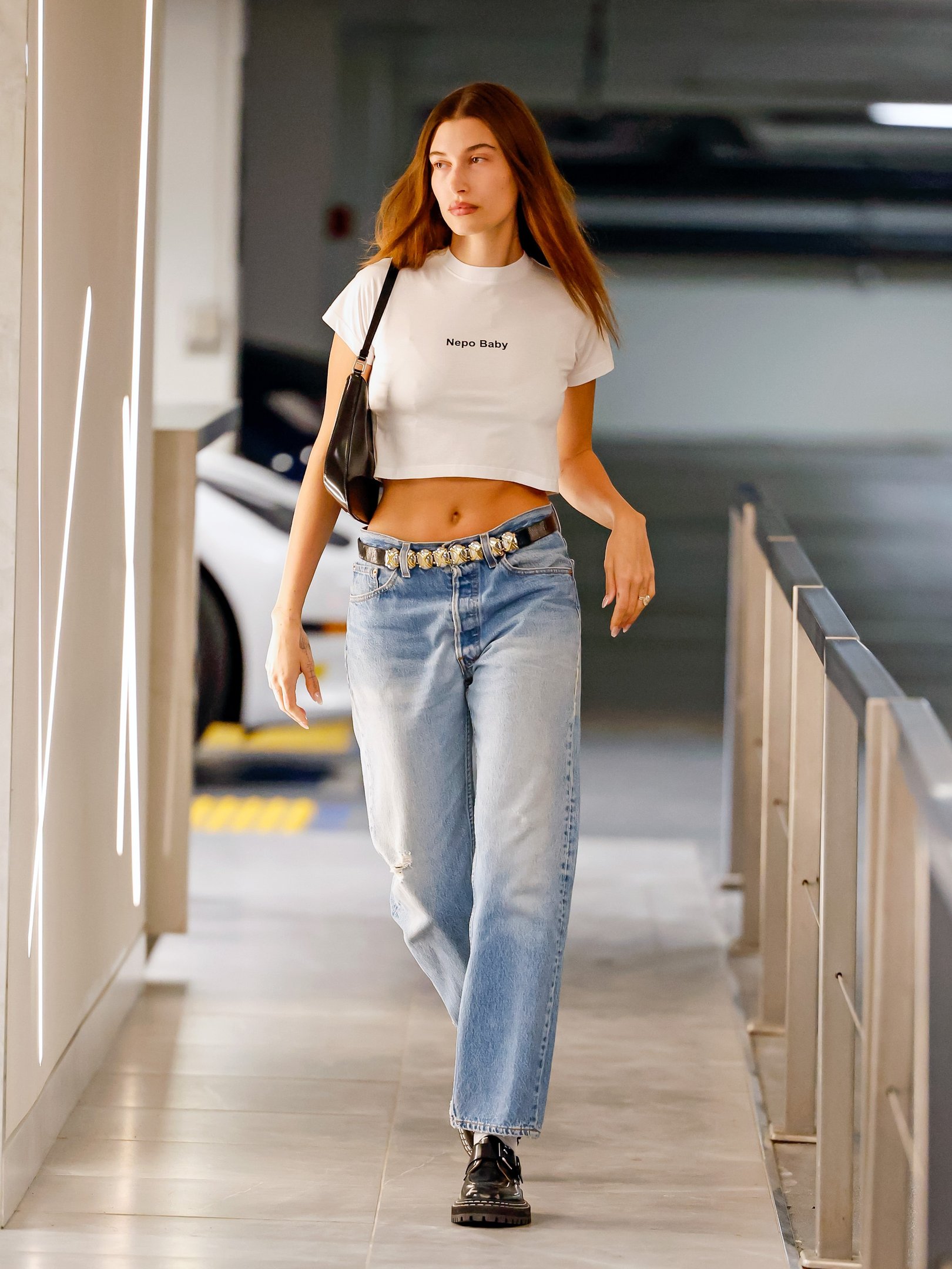 Modelo posa com camiseta cropped branca, jeans amplo azul e bolsa preta em corredor moderno. Texto: Nepo Baby