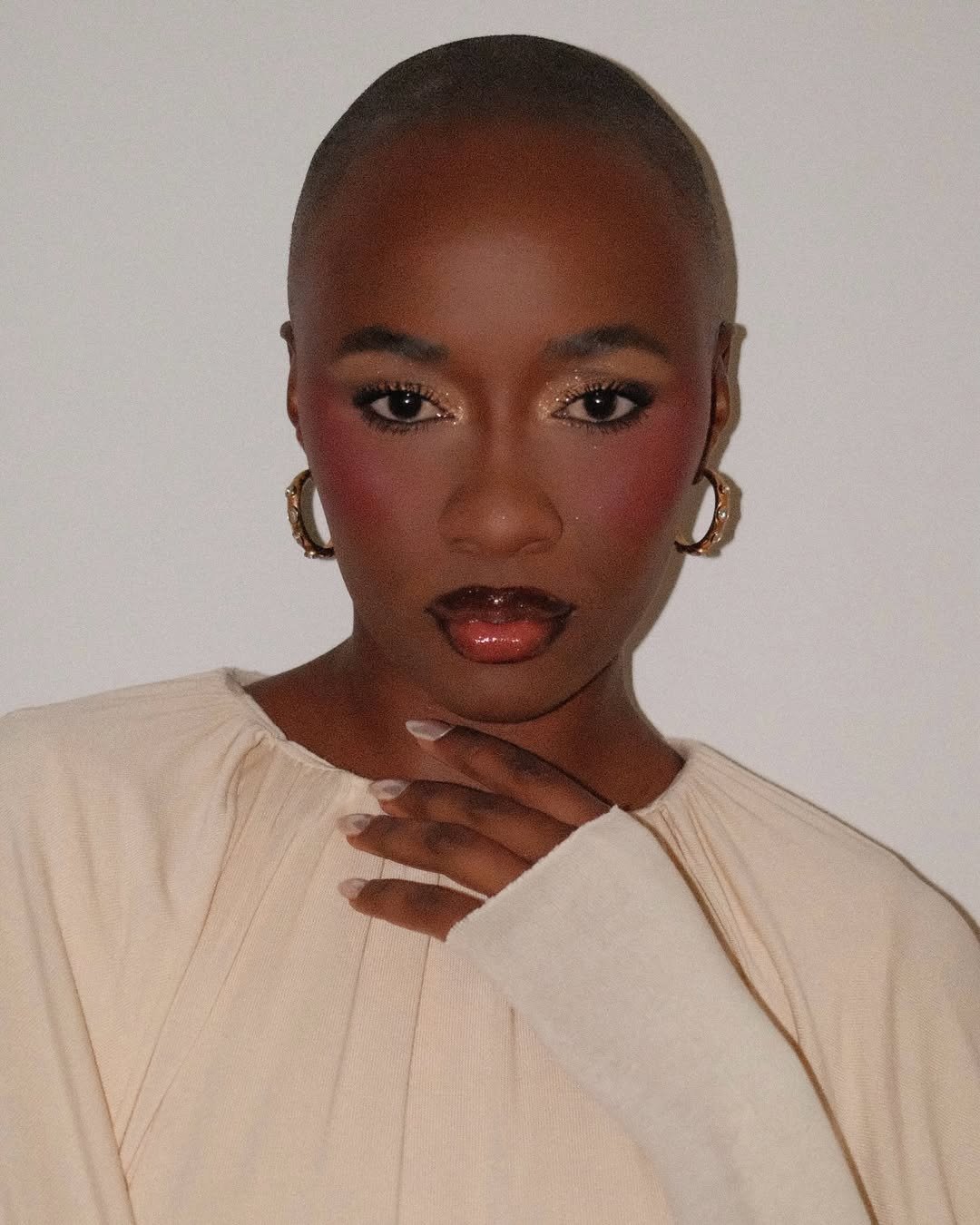 Modelo posa com blush frio rosado, sombra cintilante, argolas douradas e blusa creme ampla em clima elegante.