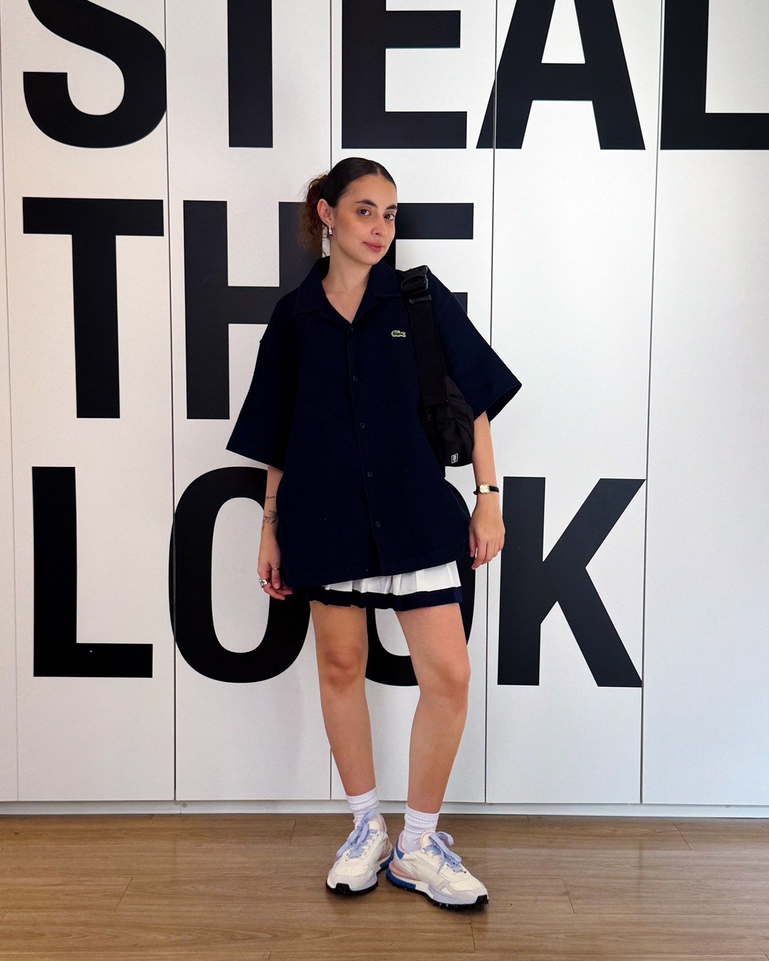 Modelo posa com tênis versátil e estiloso branco, look preto amplo e saia plissada; fundo gráfico. Texto: STEAL THE LOOK