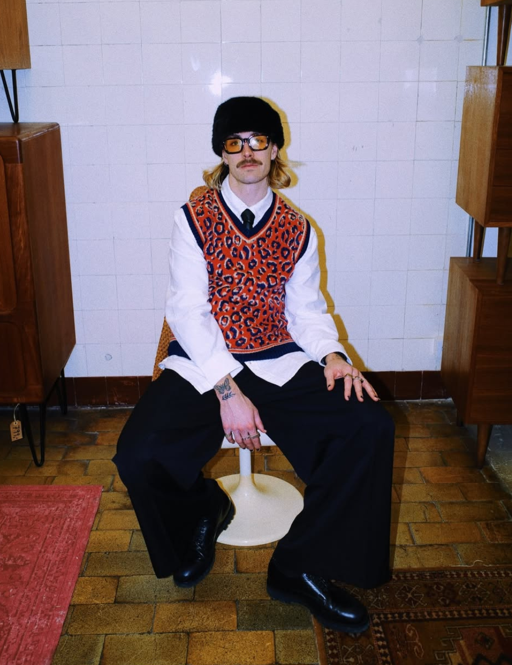 Jackson Irvine posa sentado com colete animal print laranja, camisa branca e gorro preto em clima retrô despojado.
